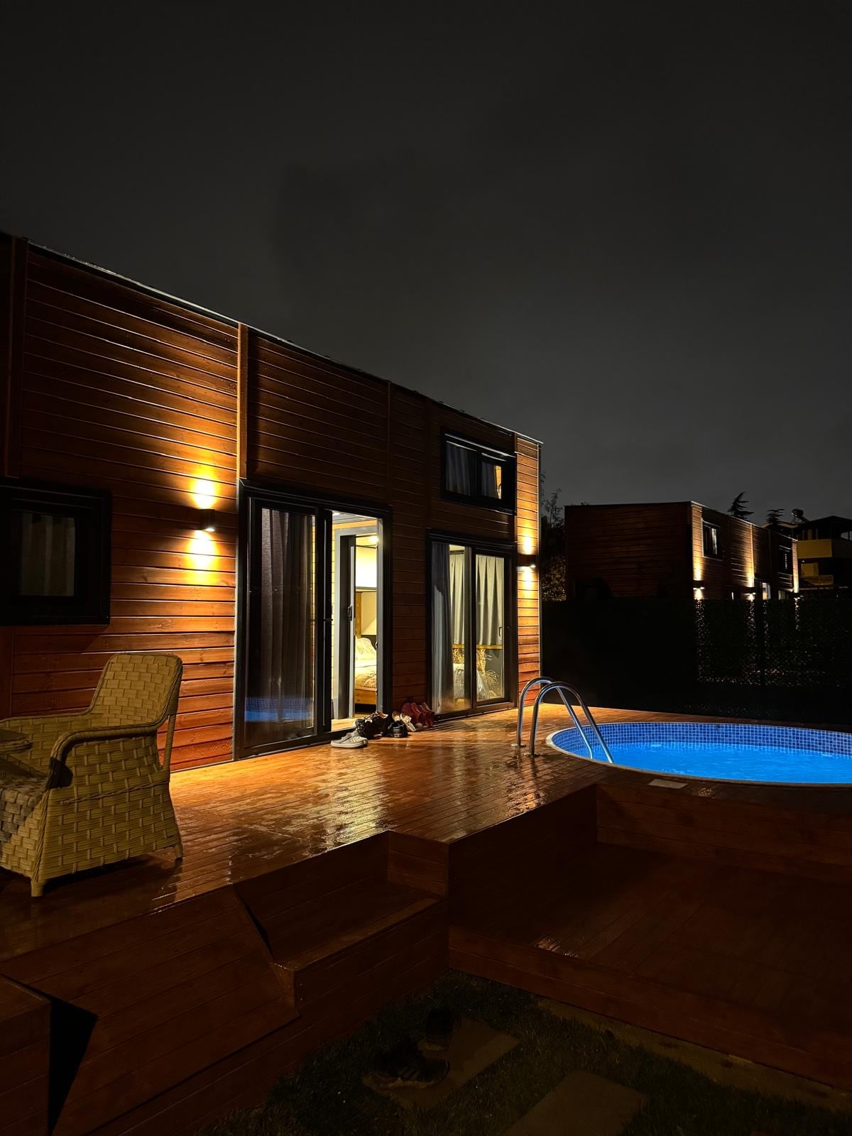 A modern tiny house is illuminated by warm outdoor lights, revealing large windows that reflect a cozy interior. A circular pool is visible on the wooden deck, while a comfortable chair adds to the inviting space. The scene is tranquil under the evening sky.
