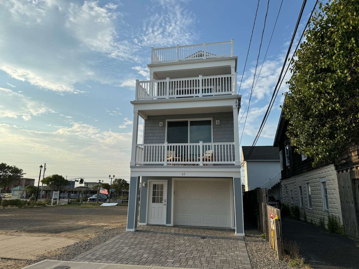A three-story new construction home features a light-colored exterior and multiple balconies. A large window at the front enhances the view of the surroundings. The driveway leads to a garage, while a well-maintained landscape surrounds the property.