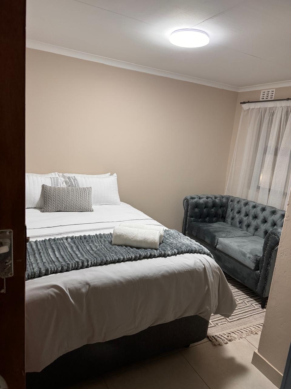 A well-appointed bedroom features a spacious bed dressed in crisp white linens. A cozy throw lies neatly across the bed. A tufted chair complements the decor, while light filters through sheer curtains near the window, creating a calm atmosphere.