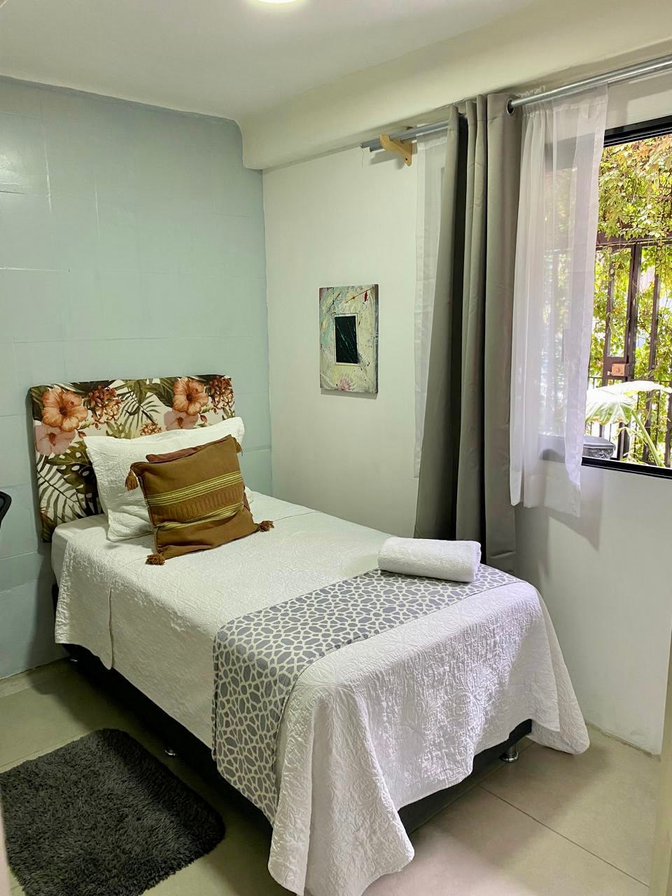 The image displays a private sleeping area with a neatly made bed, featuring floral headboard decor. Soft white linens are arranged, accompanied by a decorative accent pillow. A small towel is positioned at the foot of the bed. Natural light enters through a window with sheer curtains, revealing outdoor greenery.