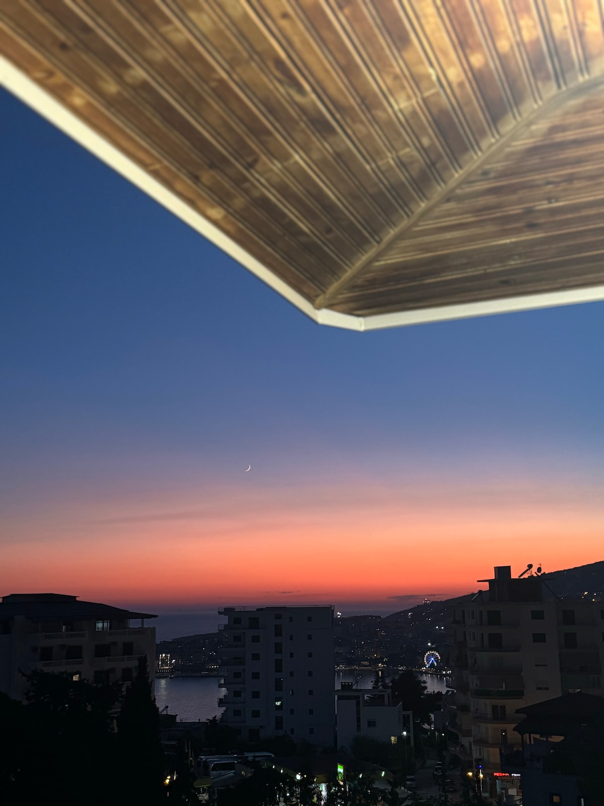 A serene sunset is captured with a gradient of warm colors transitioning from orange to purple in the sky. Silhouetted buildings are visible in the foreground, while the distant coastline reflects the twilight glow.