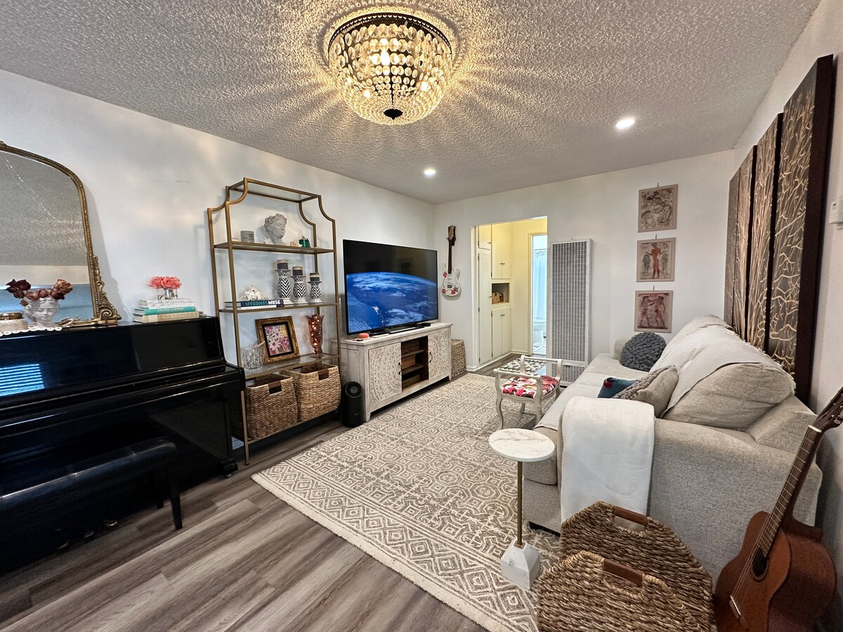 The living room presents a bright and open space with a stylish chandelier overhead. A black piano and an entertainment unit with a large television are positioned along one wall. A cozy sectional sofa faces the television, complemented by decorative accents and a textured area rug.