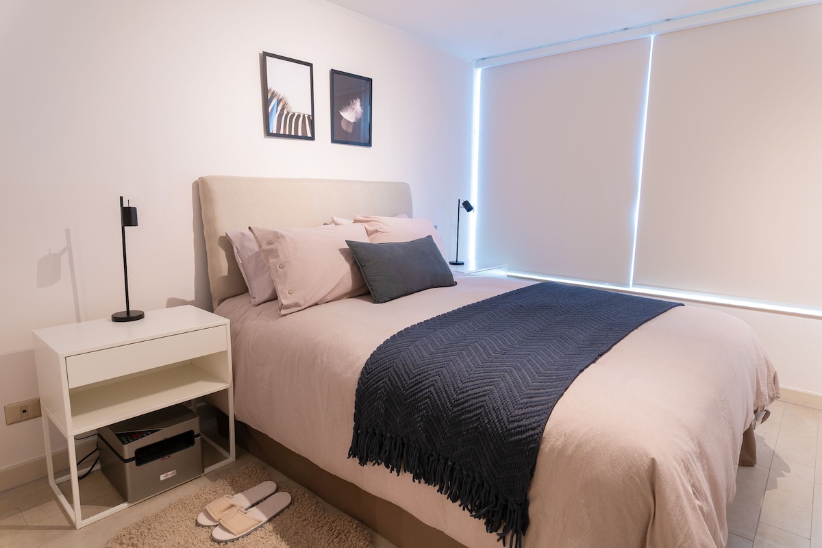 A modern bedroom features a spacious bed dressed in soft linens, with minimalistic wall art above. A nightstand with a lamp is positioned beside the bed, while natural light filters softly through the window, adorned with light-coloured roller shades.