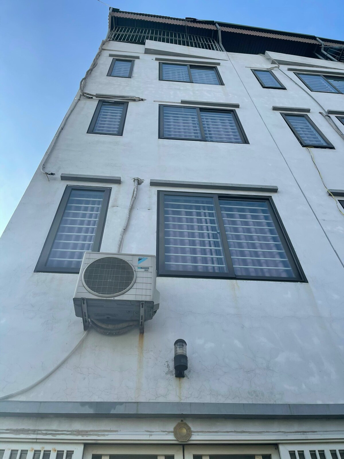 The exterior of the building is shown, featuring a clean white facade with multiple windows organized in a grid pattern. An air conditioning unit is mounted on the wall, positioned below a light fixture. The image captures the structure under a clear blue sky.