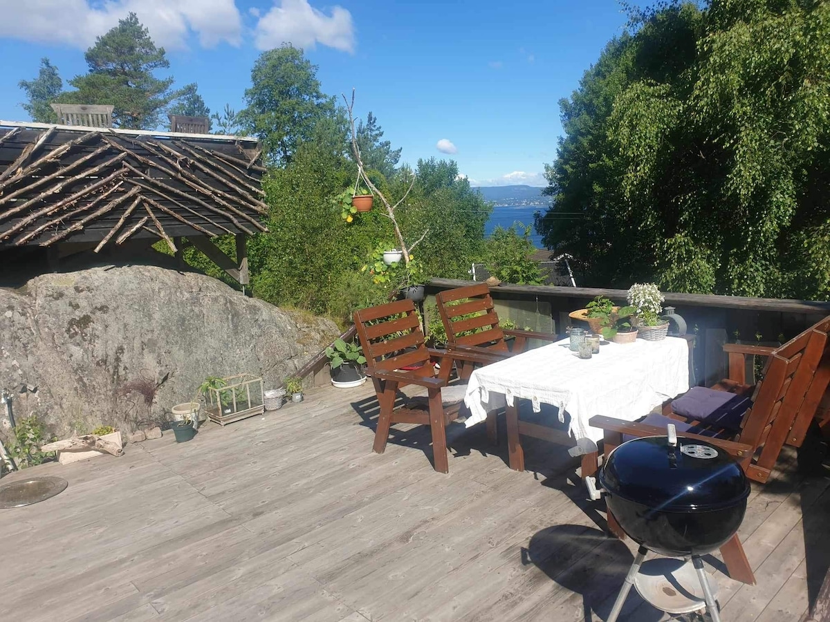 An outdoor area is displayed with a wooden deck featuring a table covered with a white cloth, accompanied by two wooden chairs. A charcoal grill stands nearby. Lush greenery surrounds the space, and a view of the sea and distant hills is visible in the background.