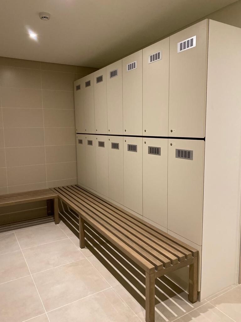 A well-organized locker area is presented, featuring multiple beige lockers with ventilation grilles. Two wooden benches provide seating against a tiled wall, offering a functional space for guests to store belongings securely.
