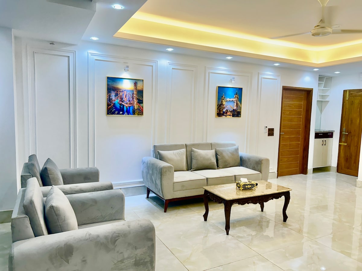 A bright living area features a light-colored sofa set and two armchairs arranged around a wooden coffee table. Soft lighting enhances the elegant wall design, while two framed artworks are displayed on the white walls. A doorway leads to a kitchen area in the background.