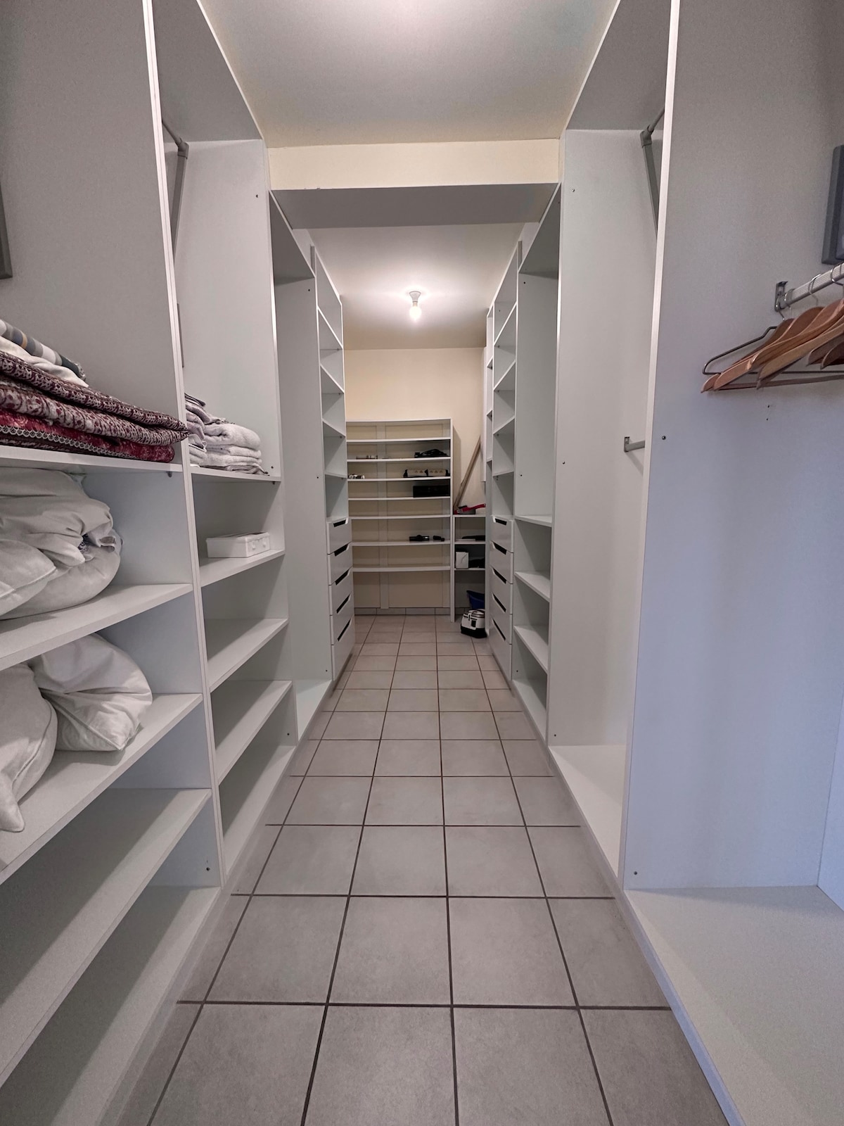 A spacious hallway features built-in shelving units on both sides, providing ample storage for linens and personal items. The floor is tiled with light-colored squares, and overhead lighting brightens the area. The design emphasizes organization and easy access to stored belongings.