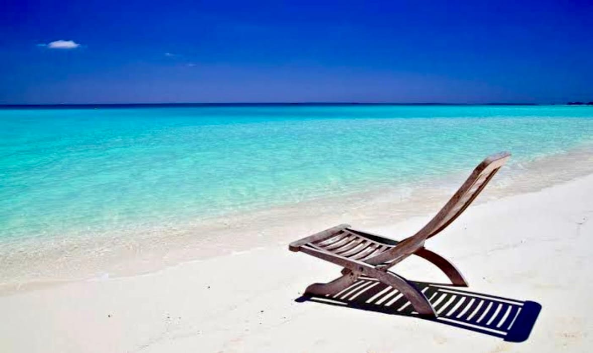 A wooden lounge chair is positioned on the sandy beach, facing the calm, clear turquoise waters. The expansive sky above is bright blue, with a few wispy clouds visible. The scene exudes a sense of relaxation and tranquility.