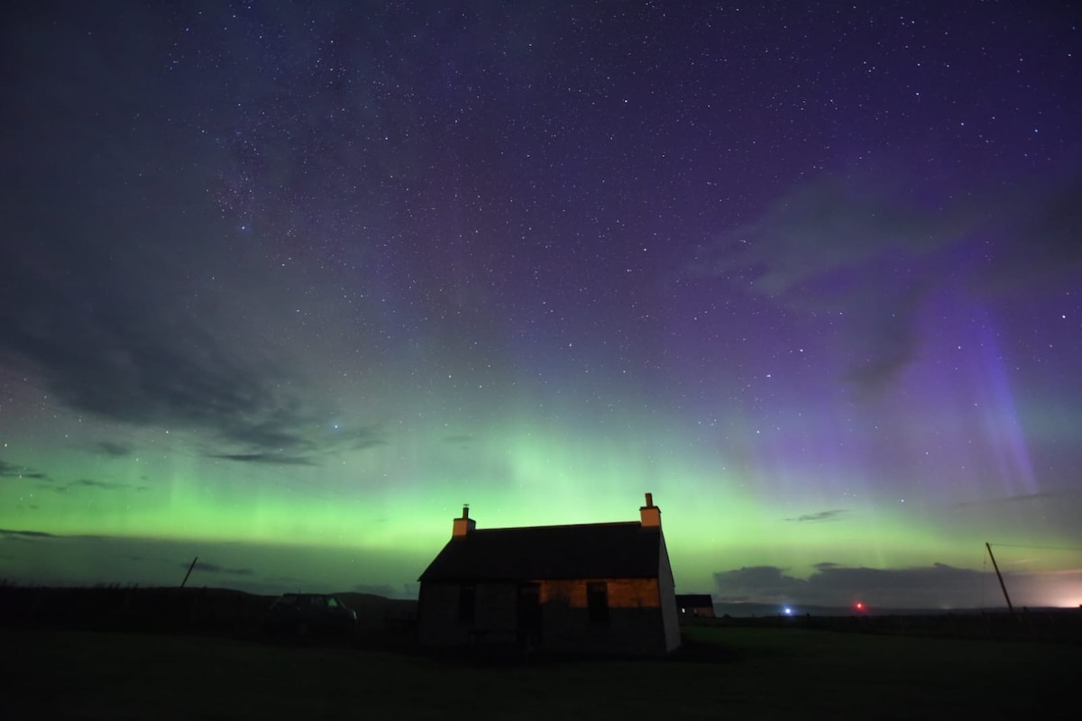 A traditional Scottish croft house is seen illuminated against a stunning night sky filled with vibrant green and purple hues of the aurora borealis. The starry atmosphere reflects the tranquility and beauty of the surrounding landscape.