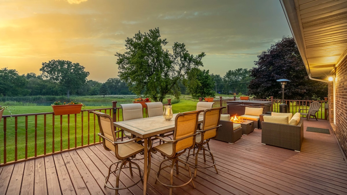 An outdoor deck is shown, furnished with a dining table surrounded by chairs and a fire pit with seating. Planters with flowers line the railing, and the lush green lawn extends toward a serene pond in the background, illuminated by a golden sunset.