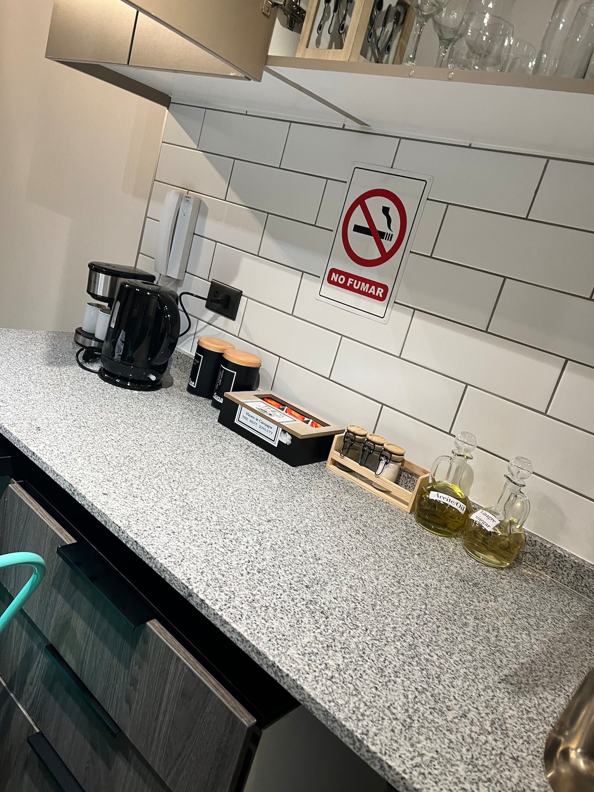 A modern kitchen countertop is set with a coffee maker, various containers, and glass bottles of oil. The sleek gray surface is complemented by a clean white tile backsplash. A sign indicating no smoking adds a safety note to the space.