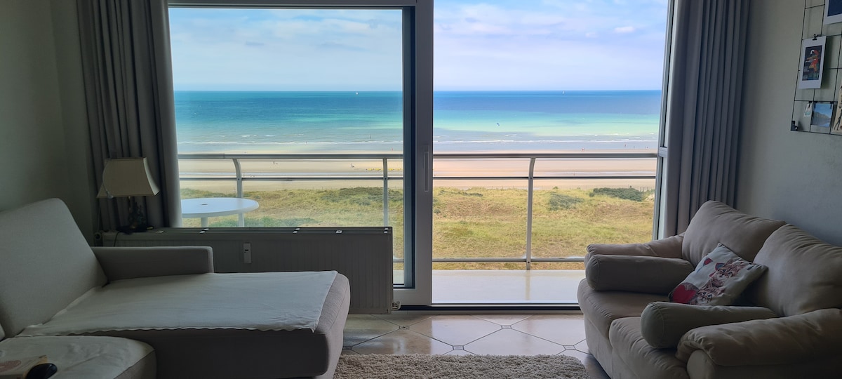 A spacious living area features large glass windows that offer unobstructed views of the sea and coastline. A comfortable sofa and long chair are arranged to take advantage of the scenic outlook, while soft natural light fills the room.