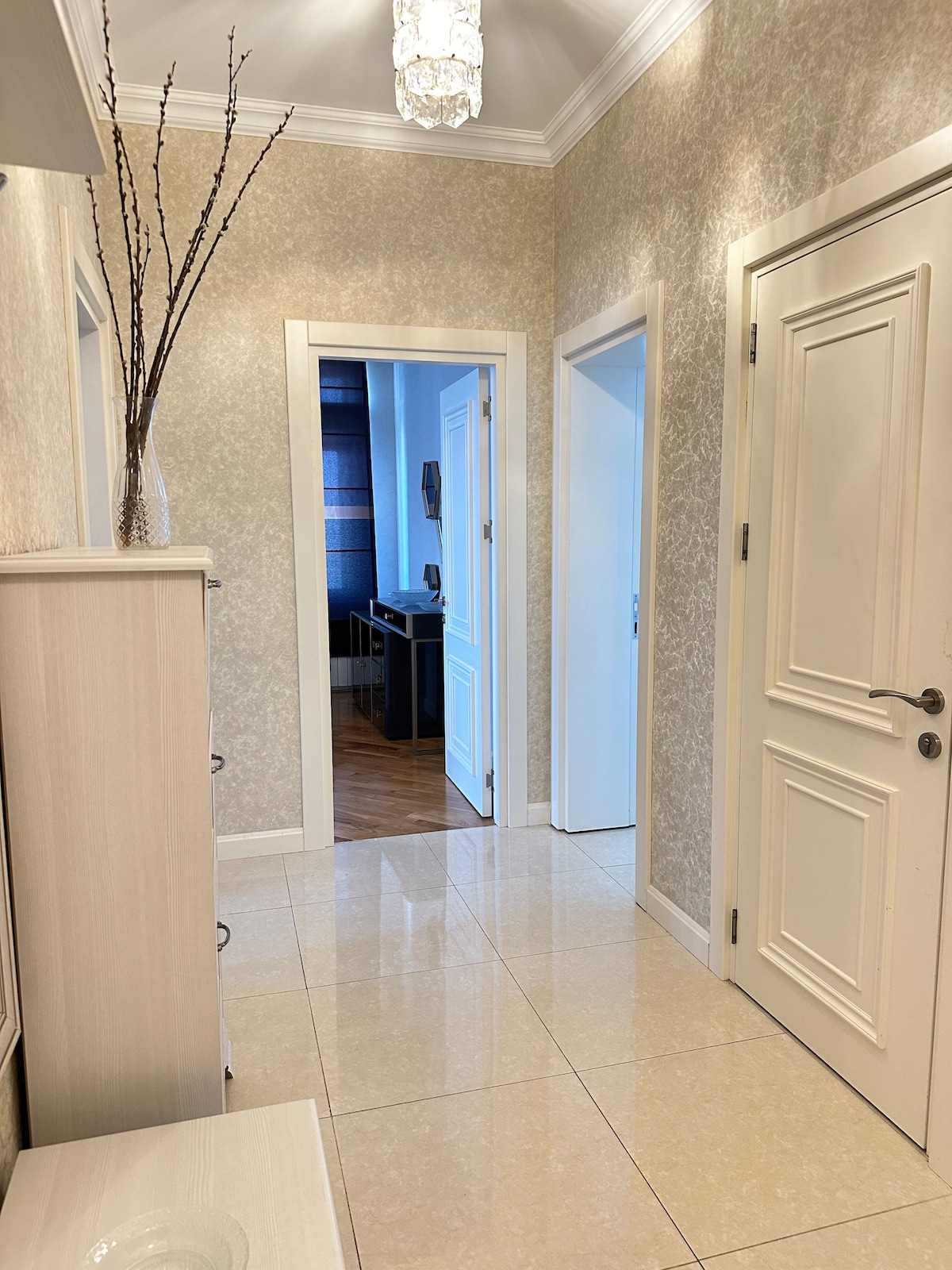 A well-lit hallway features glossy tile flooring and light-colored walls. Two doors lead off to adjacent rooms, while a small decorative vase with branches adds a touch of elegance. Soft overhead lighting illuminates the space, enhancing its welcoming atmosphere.