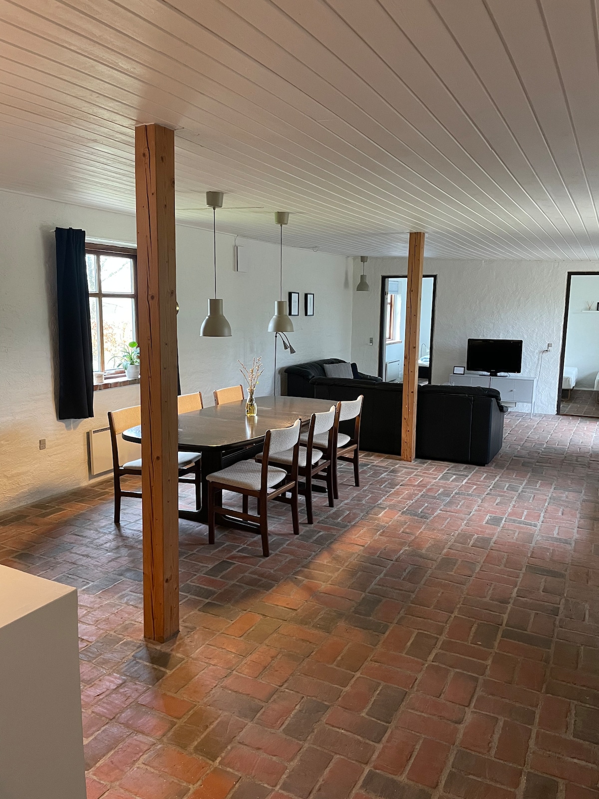 The open-plan living area features a dining table surrounded by six wooden chairs, positioned on red brick flooring. A comfortable black sofa faces a mounted television, with windows allowing natural light to brighten the space. Two pendant lights hang above the dining table.