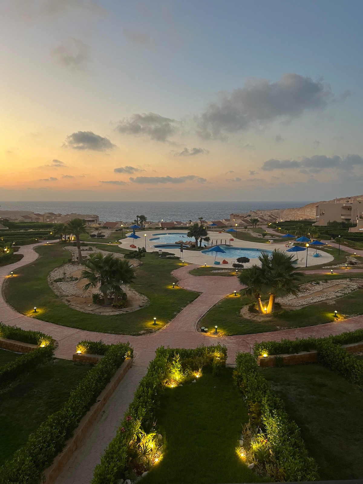 The image captures a scenic view of a landscaped area featuring winding pathways and a large swimming pool, with palm trees dotting the surroundings. The backdrop showcases a vibrant sunset over the sea, illuminating the scene with soft hues.