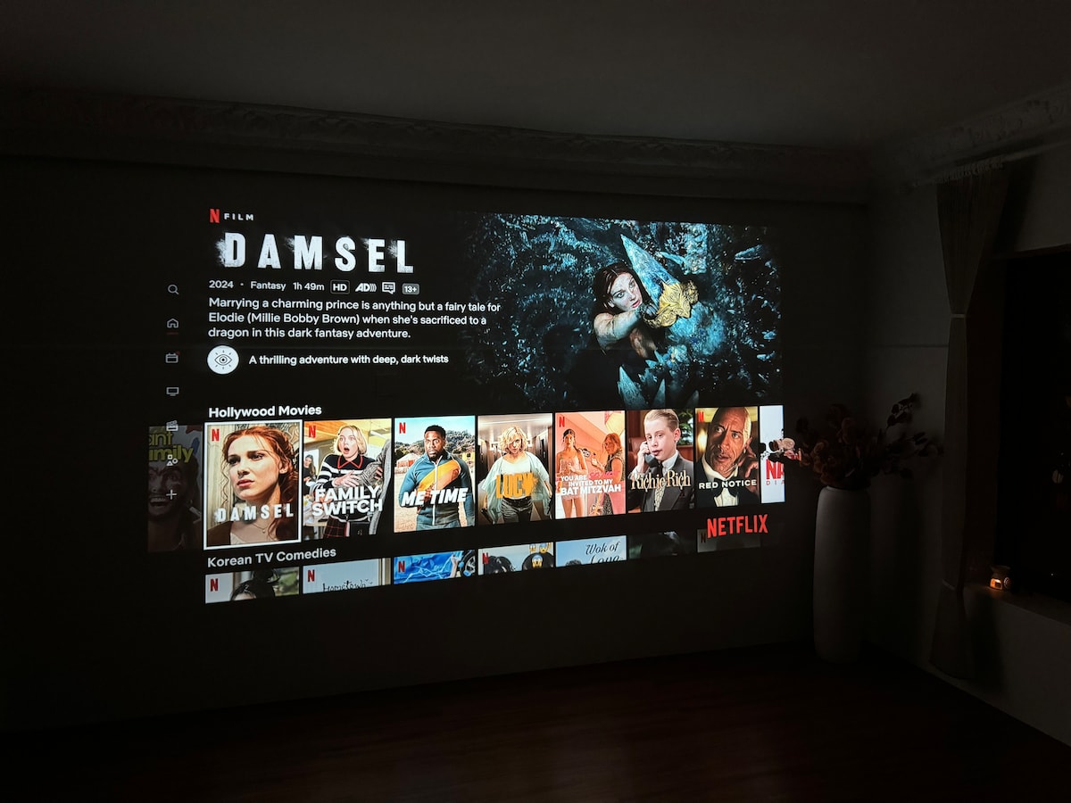 A wall is used as a screen for a 4K projector displaying the Netflix interface. The large projection features the title 'Damsel' at the top, alongside a selection of various movie thumbnails. Soft lighting from the right accentuates a decorative plant nearby.