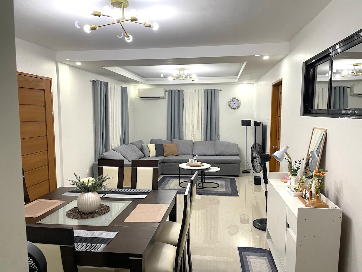 The open living and dining area features a grey sectional sofa, a glass-top dining table with seating for eight, and a stylish sideboard. Natural light fills the space through a window, complemented by light curtains and modern light fixtures.