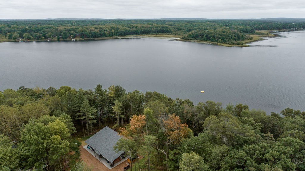 An aerial view captures a modern lake house nestled among lush trees, with a panoramic view of Benoit Lake. The calm water reflects the overcast sky, and a distant yellow swimming platform is visible in the lake, emphasizing the serene natural surroundings.