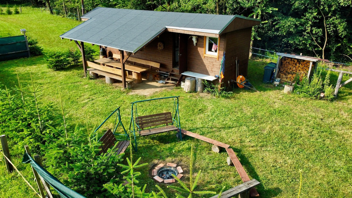 A maringotka is nestled in a serene forest setting, surrounded by lush greenery. The outdoor area features a fire pit bordered by wooden seating and a spacious deck. A storage area and various trees enhance the inviting environment, showcasing a peaceful escape in nature.