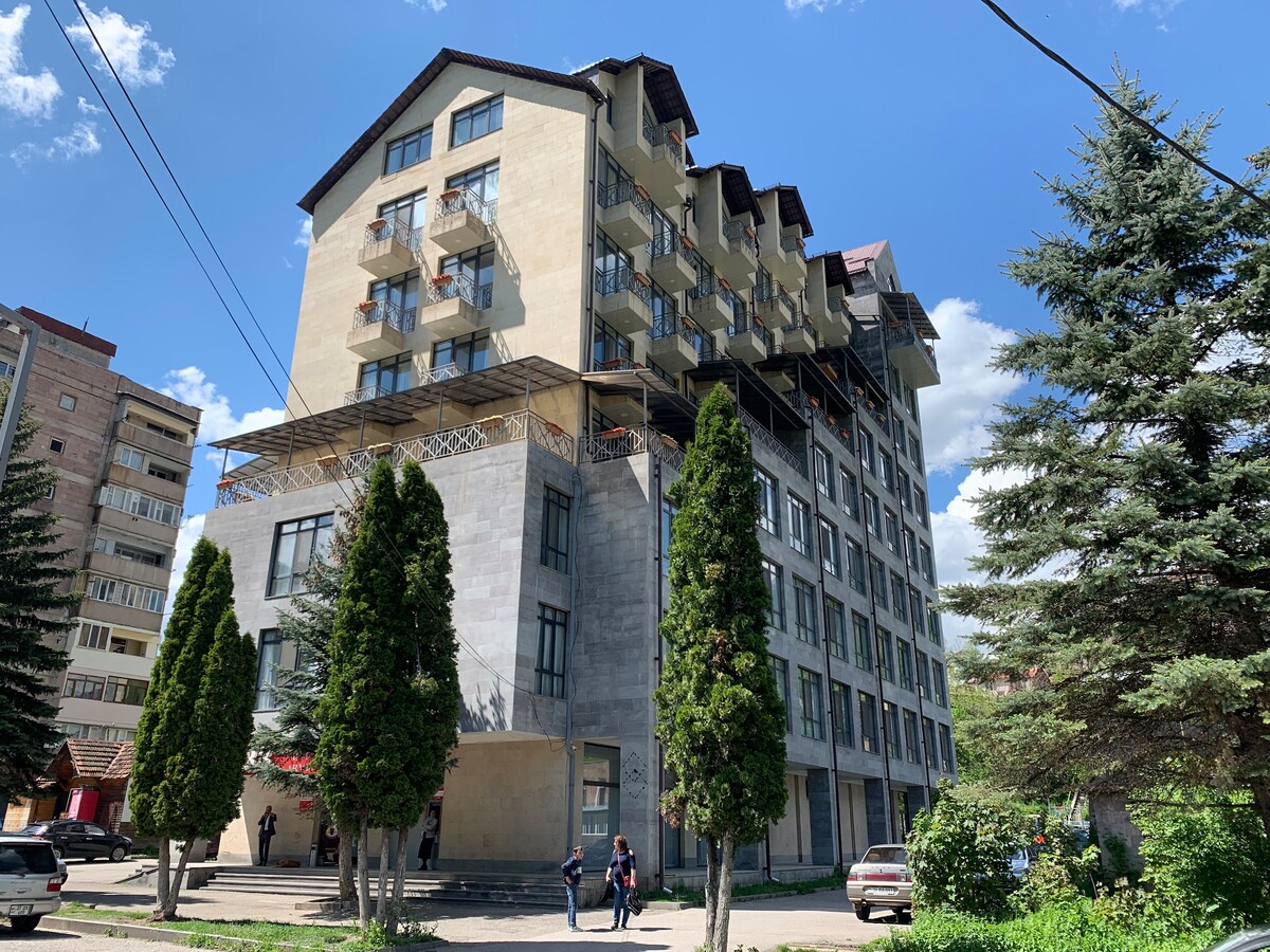 A multi-story building features a mix of concrete and glass facades. Balconies are visible on each level, providing outdoor space. Tall trees line the front, offering greenery against the backdrop of a clear blue sky.