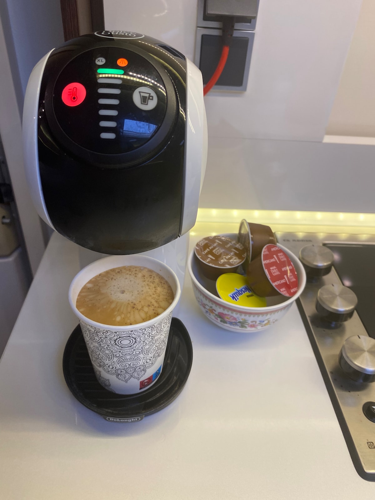 A coffee machine is positioned on a sleek countertop, producing a cup of coffee placed on a drip tray. A bowl containing coffee capsules and creamers is situated nearby, while stainless steel knobs of a stove are visible in the background.