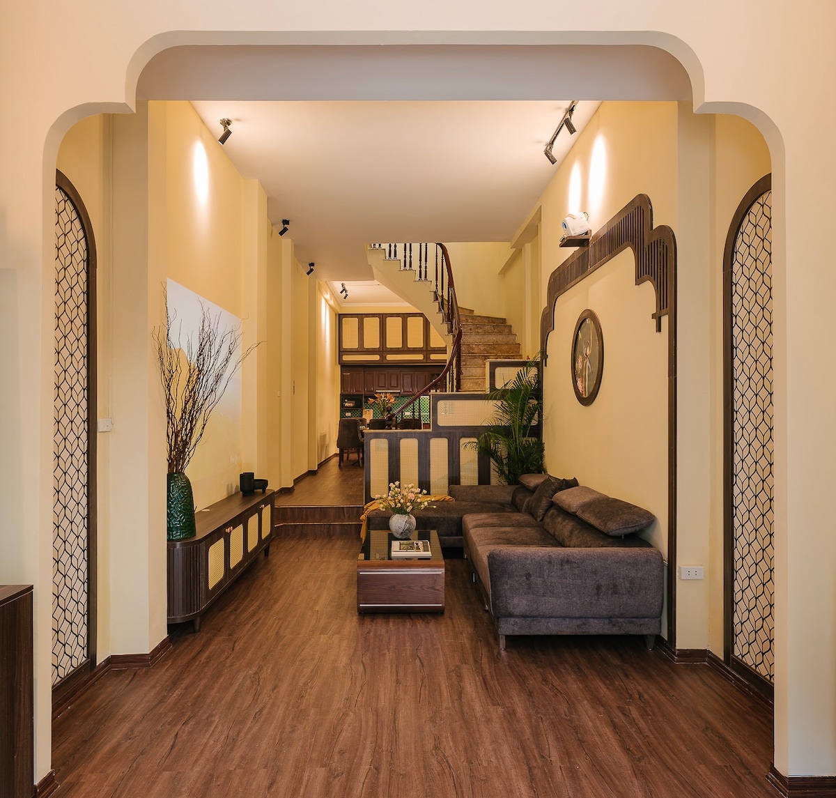 A welcoming living area features a spacious, L-shaped sofa facing a decorative coffee table. Natural light is provided by ceiling fixtures and wall sconces. The open layout leads towards a staircase and a partially visible kitchen area, enhancing the home's inviting atmosphere.