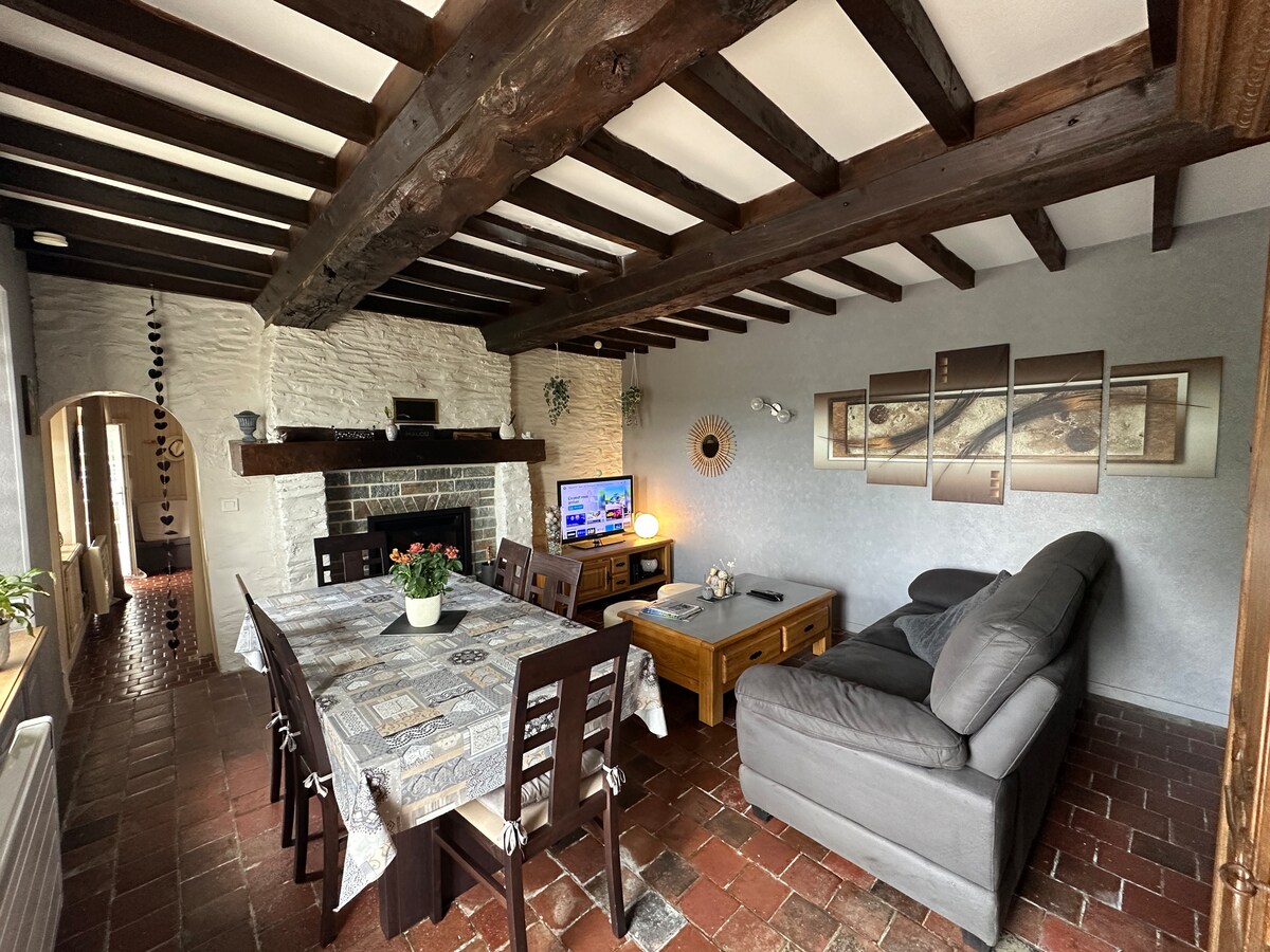 A lounge area showcases a comfortable gray sofa and a wooden coffee table. A dining table with seating for six complements the space. An extensive wooden beam ceiling adds character, while a flat-screen television is positioned on a console below a wall art display.