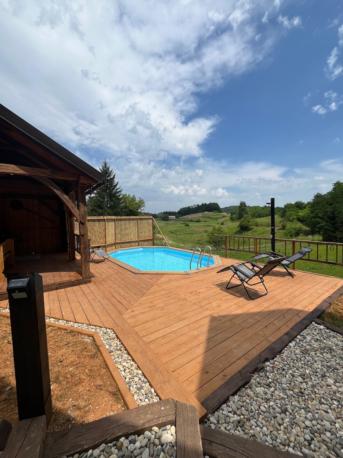 An outdoor area features a wooden deck surrounding a bright blue swimming pool. Two lounge chairs are positioned beside the pool, offering a relaxing place to enjoy the scenic views of the green landscape under a partly cloudy sky.
