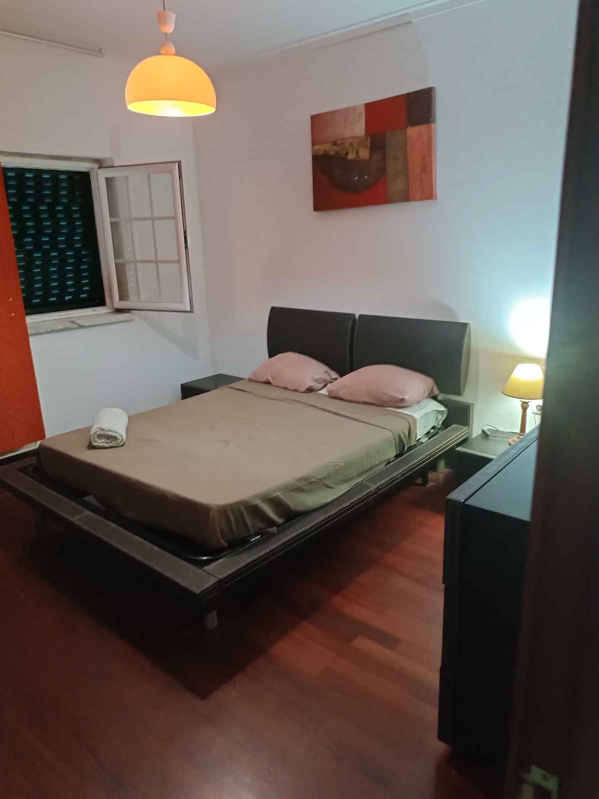A cozy sleeping area is presented with a spacious bed featuring a simple beige bedspread. A bedside lamp provides gentle lighting, and a colorful wall art piece hangs above the bed. A window with partially open shutters allows some natural light into the room.