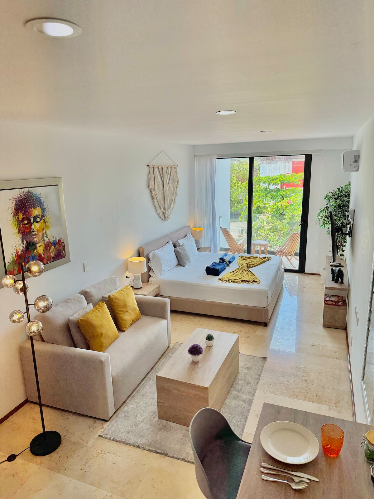 A bright studio space showcases a spacious bed, complemented by a light-colored sofa adorned with decorative cushions. A small dining area is visible, set with tableware. Large glass doors provide access to an outdoor area with greenery, enhancing the open feel of the room.