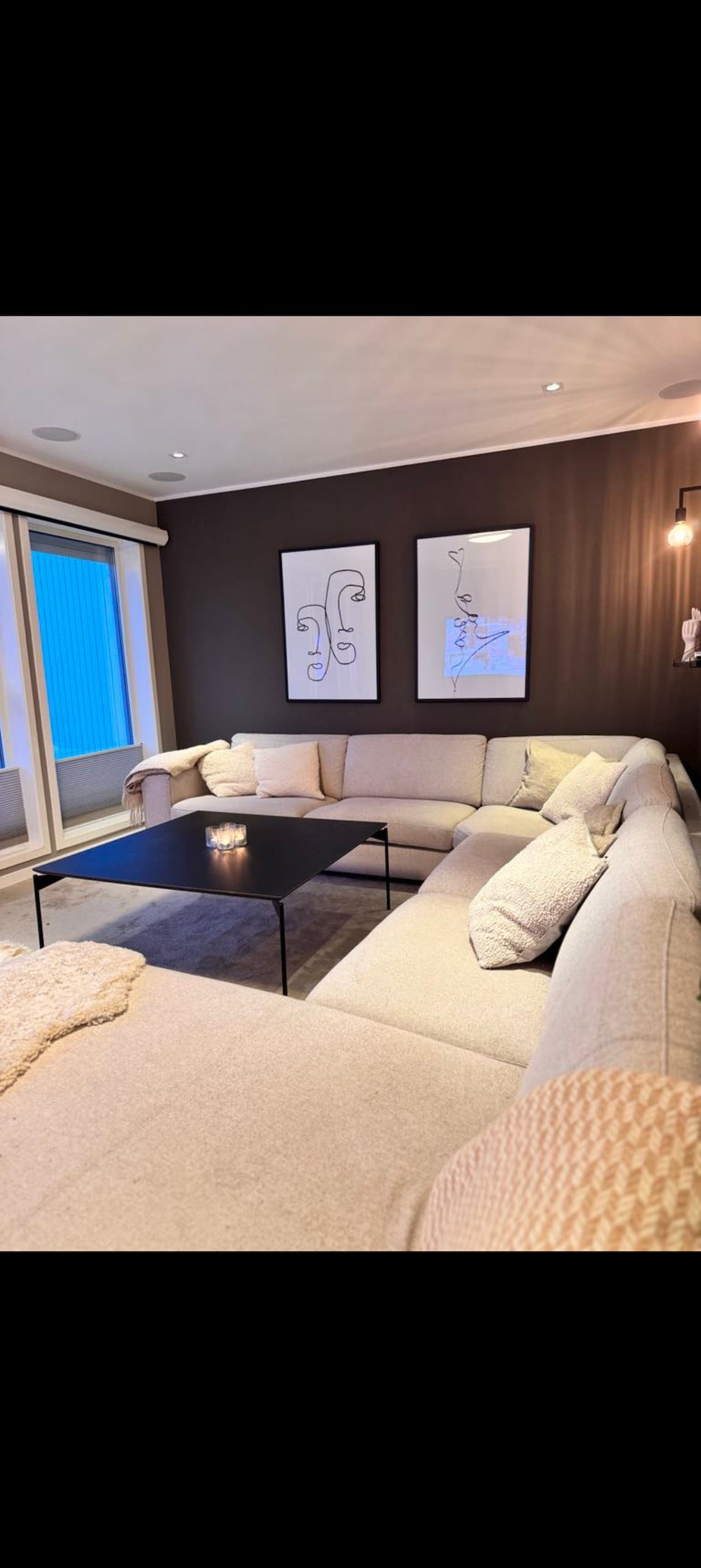 A spacious living area features a large sectional sofa, arranged around a sleek black coffee table. Two framed artwork pieces adorn the dark walls, and natural light enters through large windows, creating a cozy environment for relaxation and gatherings.