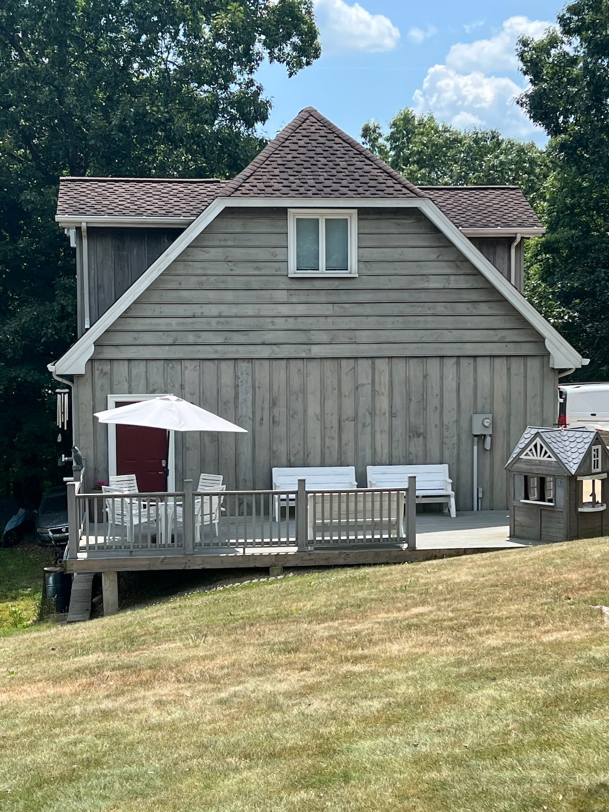 The exterior of the two-story wooden structure is shown, featuring a classic gabled roof and a large deck. A shaded umbrella stands over a seating area that includes white chairs, while a small playhouse adds charm to the surrounding yard.
