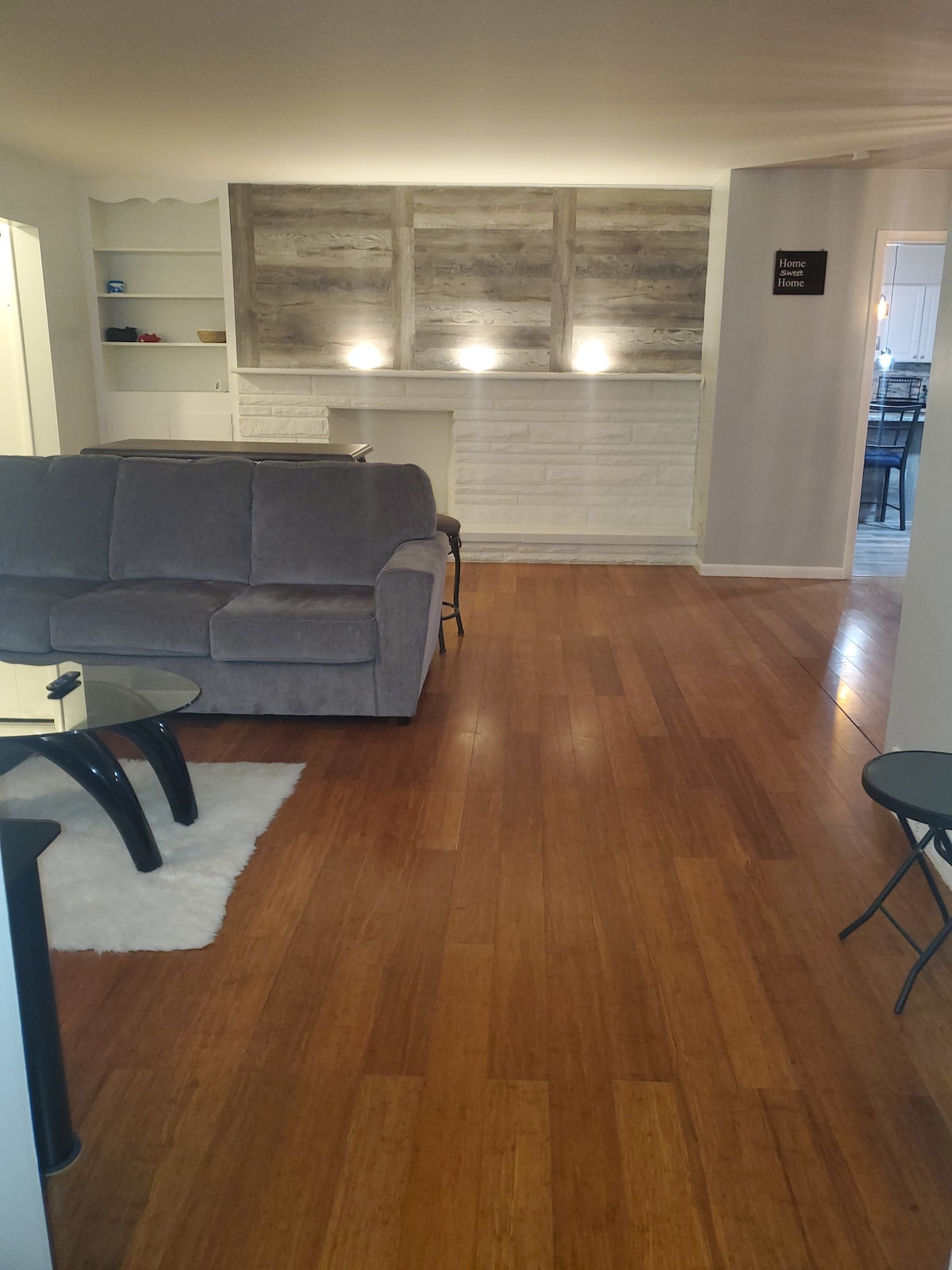 A spacious living area features a comfortable gray couch positioned facing a glass coffee table on a soft white rug. Light illuminates the textured wall behind the couch, enhancing the hardwood flooring throughout the space.