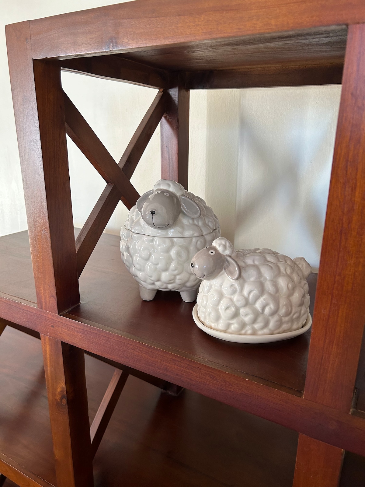 Two ceramic sheep figurines are displayed on a wooden shelf. The sheep are textured to resemble wool, with a soft grey hue. The shelf is made of dark wood, featuring a cross design in the side panels, creating a simple yet charming decorative element.
