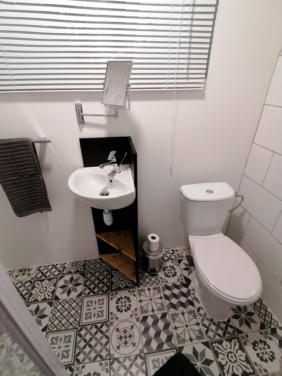 A modern bathroom is displayed, featuring a white sink with a sleek faucet mounted on a black shelf. A white toilet is positioned beside it. Textured tiles with geometric patterns cover the floor, while a mirror hangs above the sink. Natural light filters through the window covered with blinds.