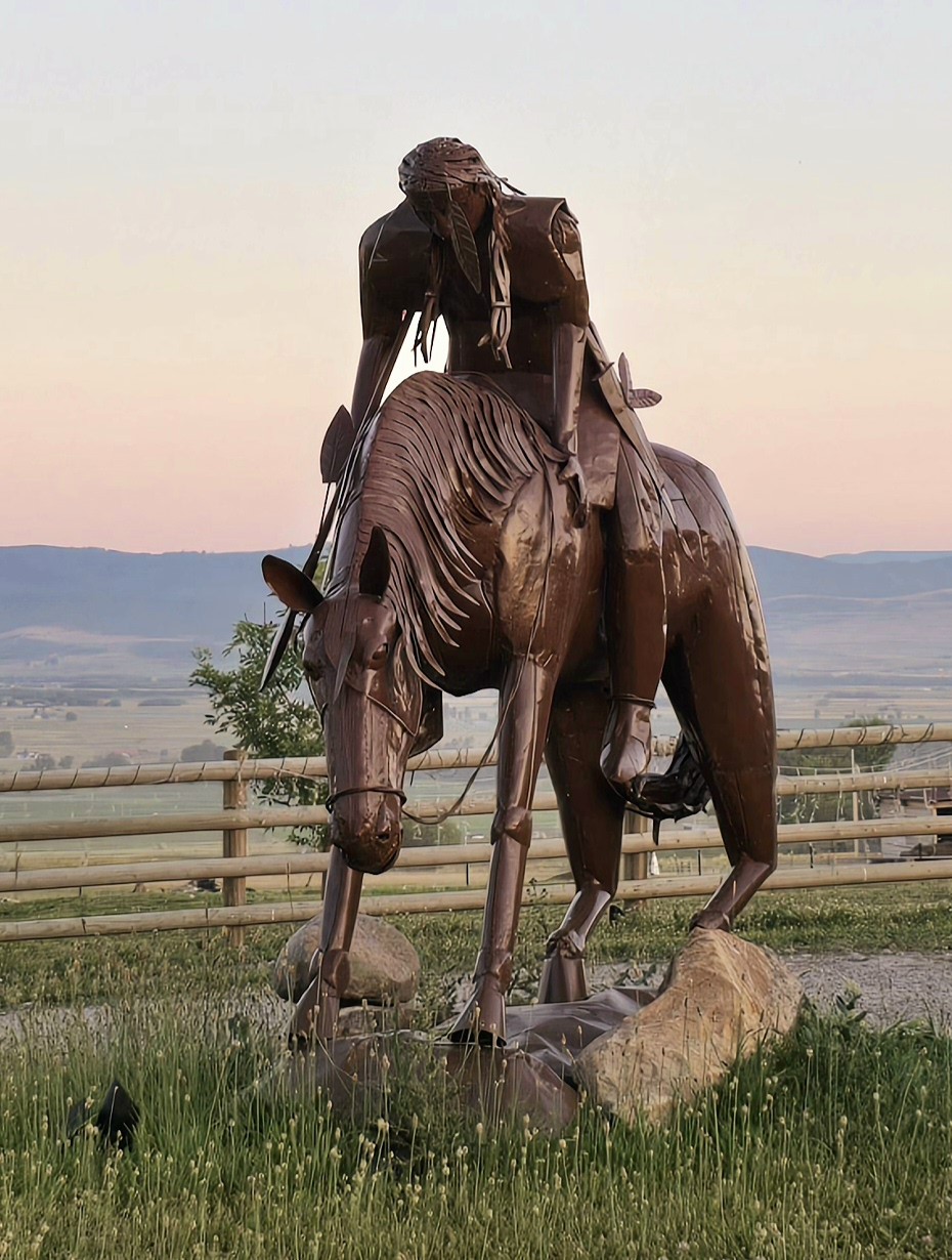 A metal sculpture of a horse and rider is positioned in a grassy area. The rider appears to be leaning forward gently, while the horse stands elegantly on a stone base. A soft gradient of pink and purple sky is visible in the background.