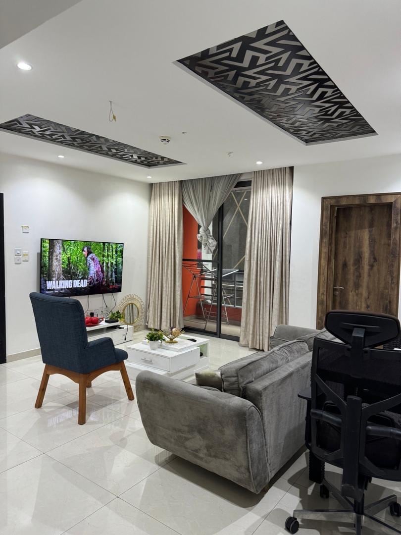 A modern living room is displayed, featuring a comfortable grey sofa and a blue accent chair. A flat-screen TV is mounted on the wall, showcasing a nature scene. Light-colored curtains frame the window, while a coffee table with decor sits in the center. A desk chair is positioned nearby.
