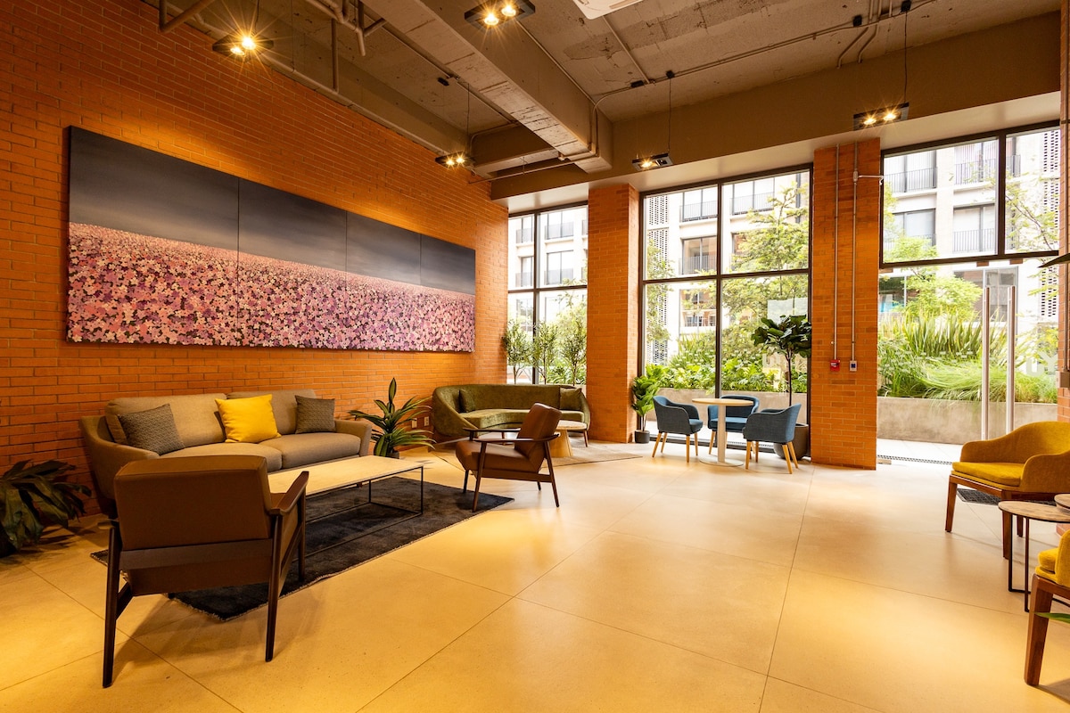 A communal lounge area features large windows that fill the space with natural light. Modern furniture, including a sectional sofa and armchairs, is arranged around a low coffee table. A large floral artwork hangs on the wall, complementing the warm brick accents.