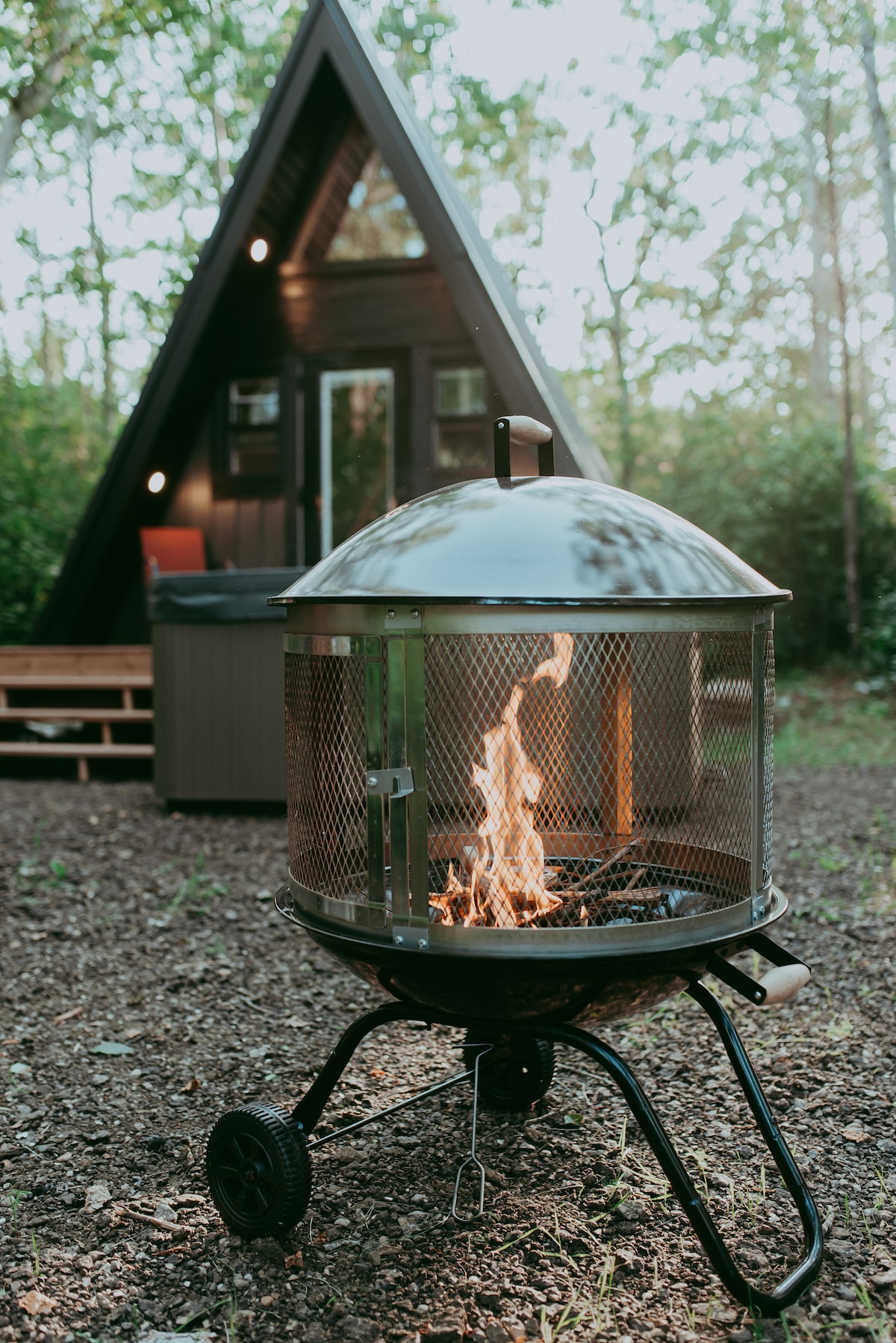 A fire pit with a metallic mesh lid is positioned on a gravel surface in front of a cozy A-Frame cabin. Flickering flames are visible within the fire pit, surrounded by a serene wooded area, enhancing the outdoor ambiance.
