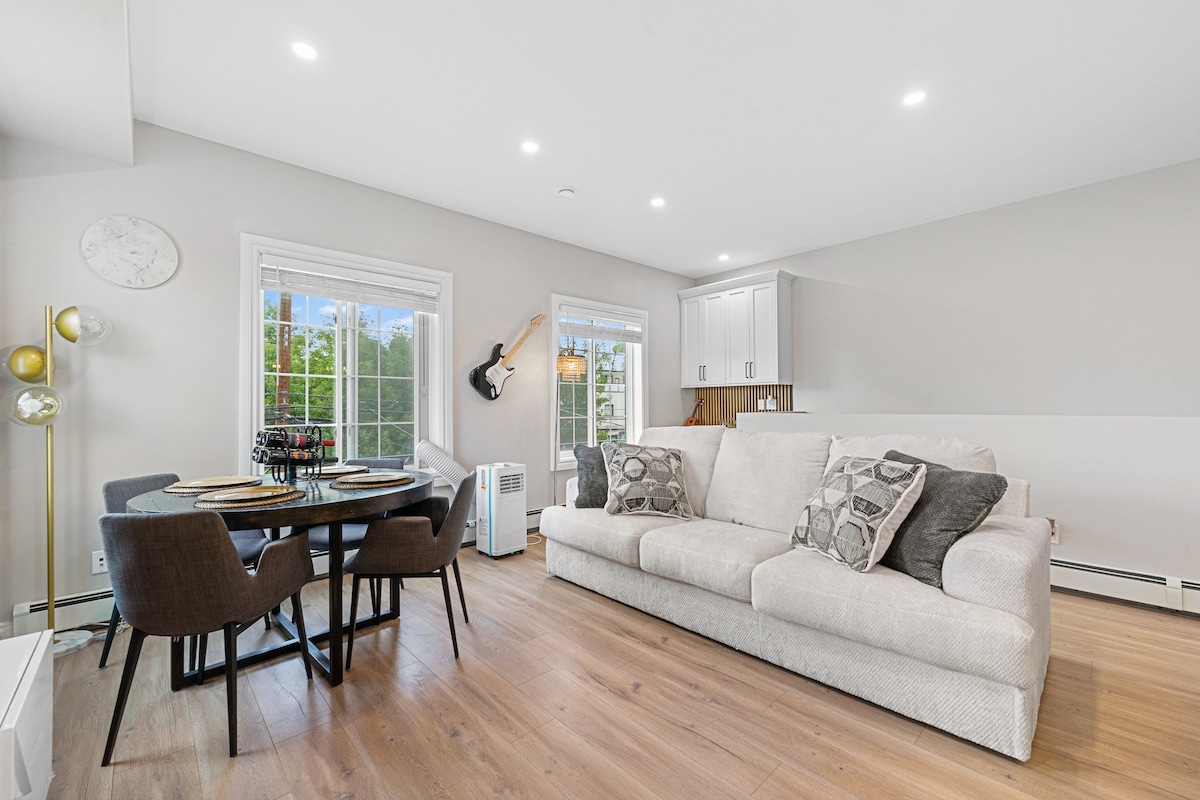 A modern living area features a spacious beige sofa adorned with patterned cushions. A round dining table with dark chairs is positioned nearby, with natural light streaming in through large windows. A guitar is mounted on the wall, adding a touch of character to the space.