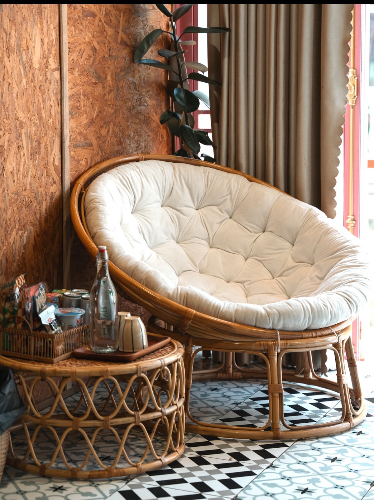 A round, cushioned chair made of wicker is positioned against a textured wall. A small, round side table made of rattan holds a bottle of water and a decorative basket. Nearby, a leafy plant is seen, and patterned floor tiles contrast with the warm tones of the seating area.