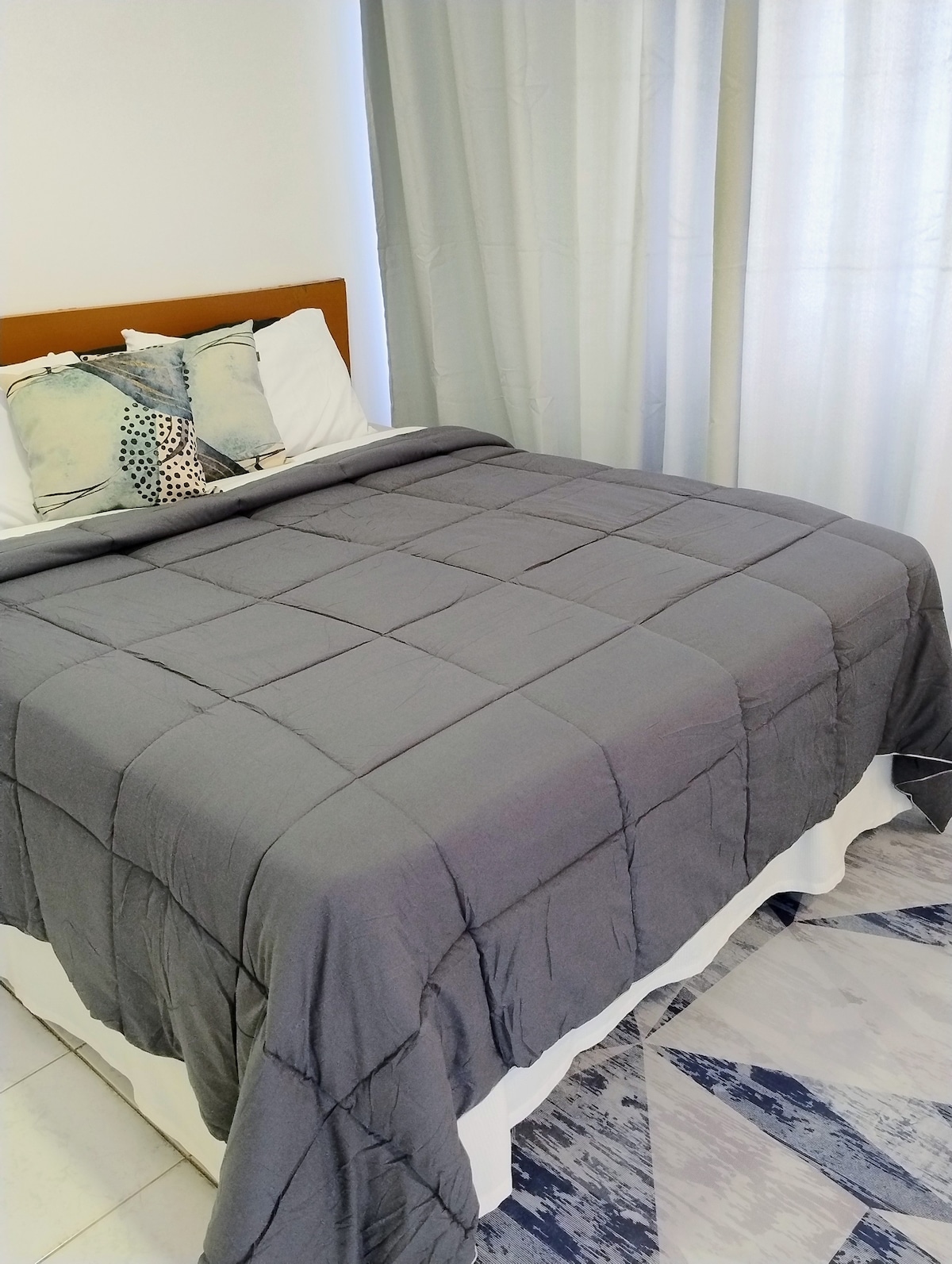 A spacious bed is dressed in a grey quilt, complemented by decorative pillows. Soft light filters through sheer curtains, illuminating the linen and adding a calming ambiance. The floor features light-colored tiles, creating a clean and airy environment.