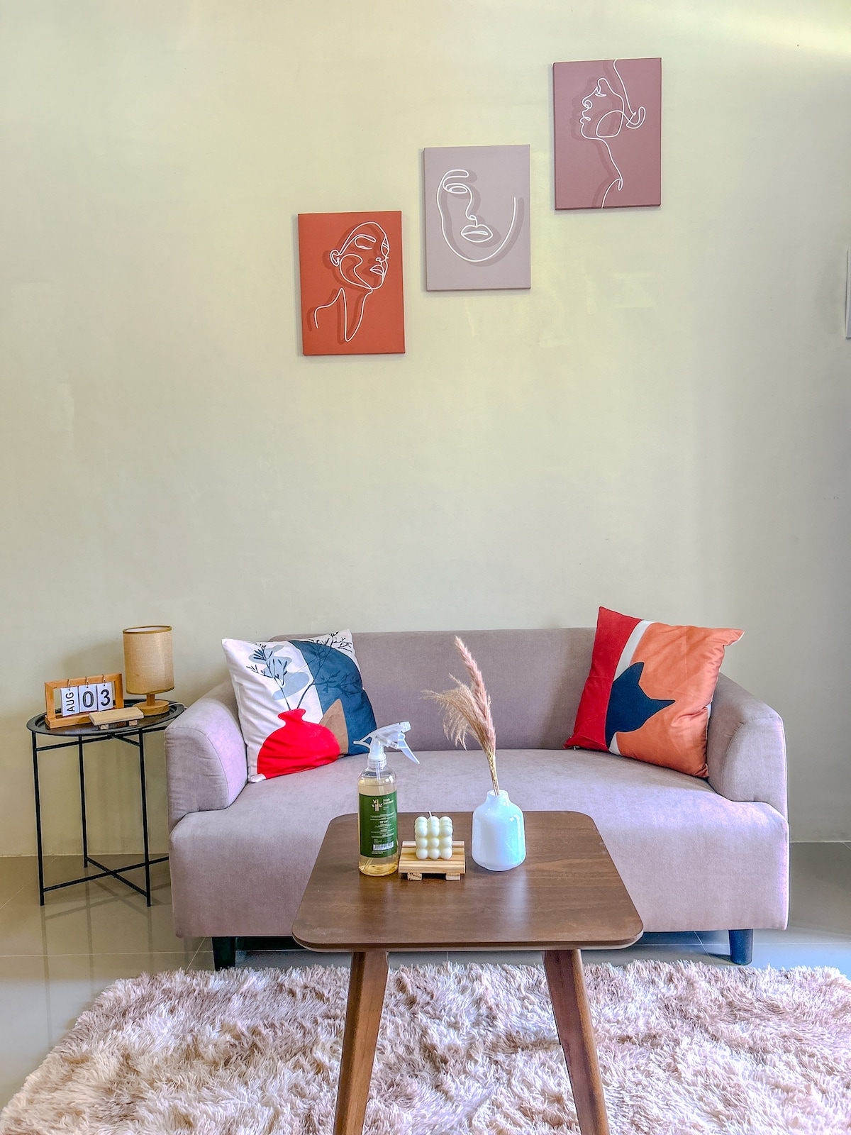 A minimalist living area is presented with a soft beige couch adorned with colorful cushions. A small wooden coffee table is positioned in front, featuring decorative items. Three line-art portraits hang above the couch, contributing to a serene atmosphere.