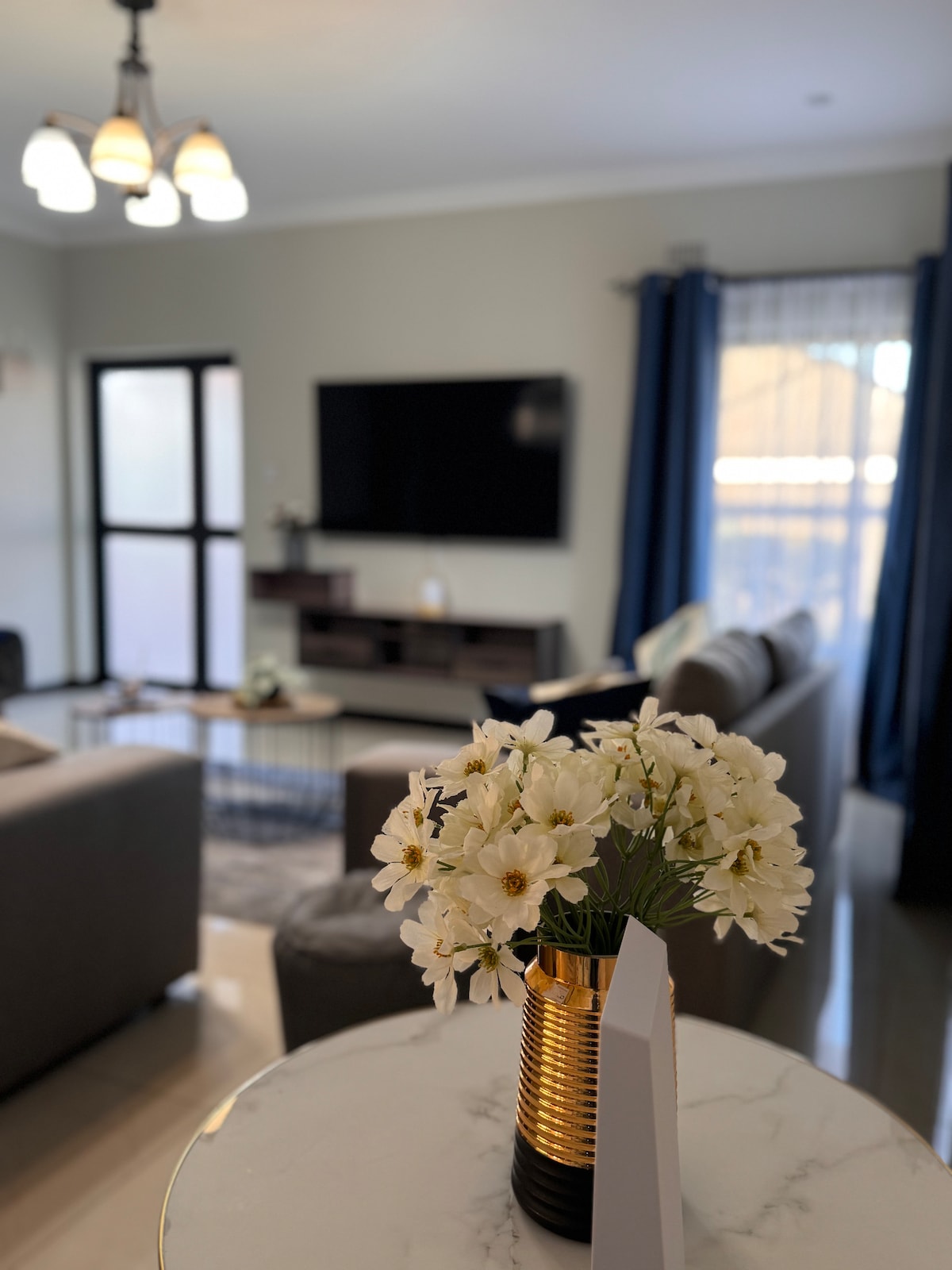 A living area is featured with comfortable seating arranged around a coffee table. A television is mounted on the wall, providing entertainment options. A vase with white flowers sits on a marble surface, complementing the natural light flowing through nearby windows with curtains.