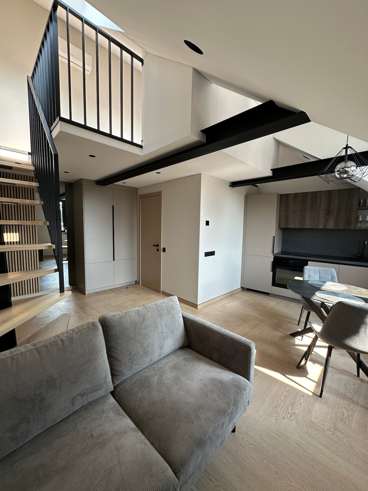 A modern living space is showcased, featuring a light-colored sofa positioned in the foreground. The open layout reveals a staircase leading to the upper level, where a balcony can be seen. A dining table with chairs is situated near the kitchen area.
