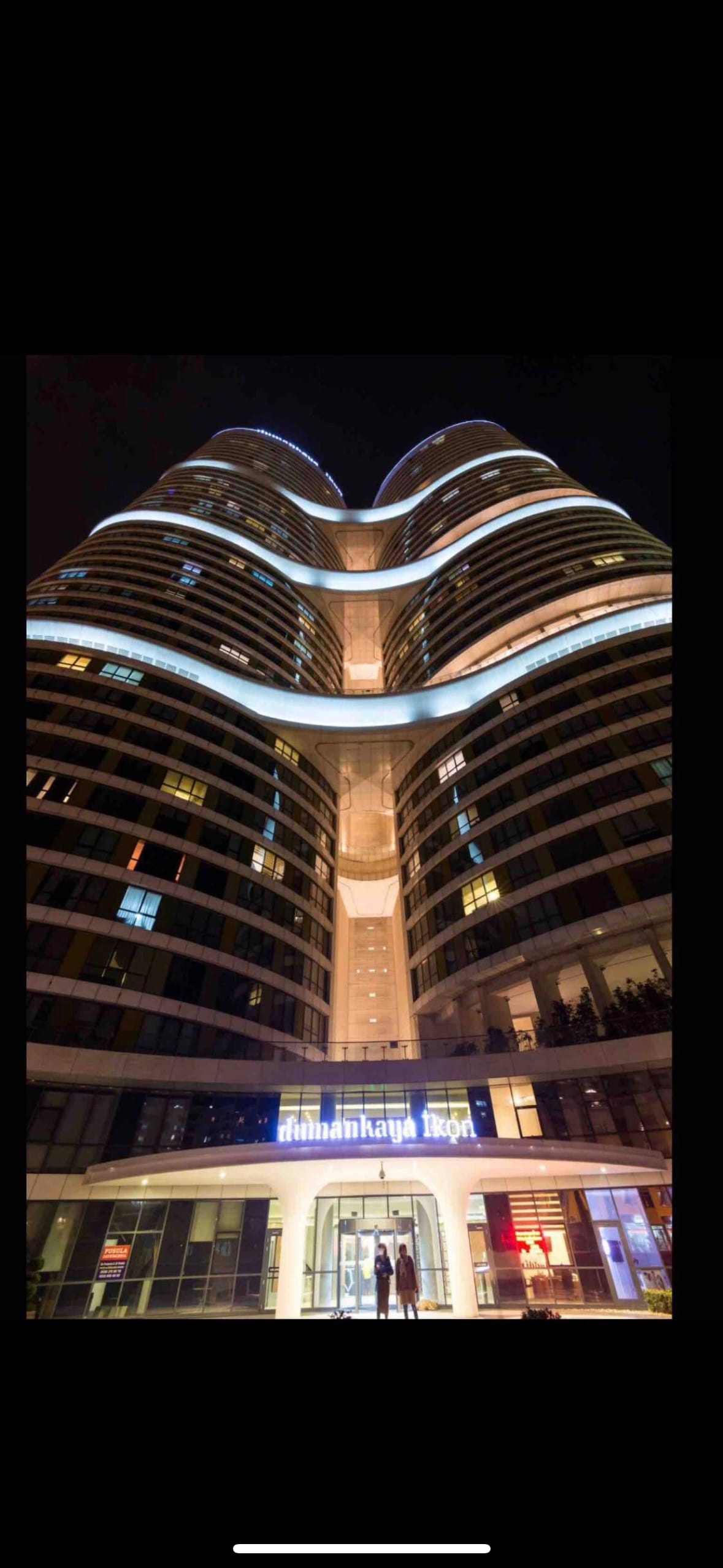 The exterior of a modern high-rise building is illuminated at night, showcasing sleek lines and a unique architectural design. Bright lights reflect in the windows, emphasizing the dual curves of the structure. The entrance is marked by a welcoming sign, adding a professional touch.