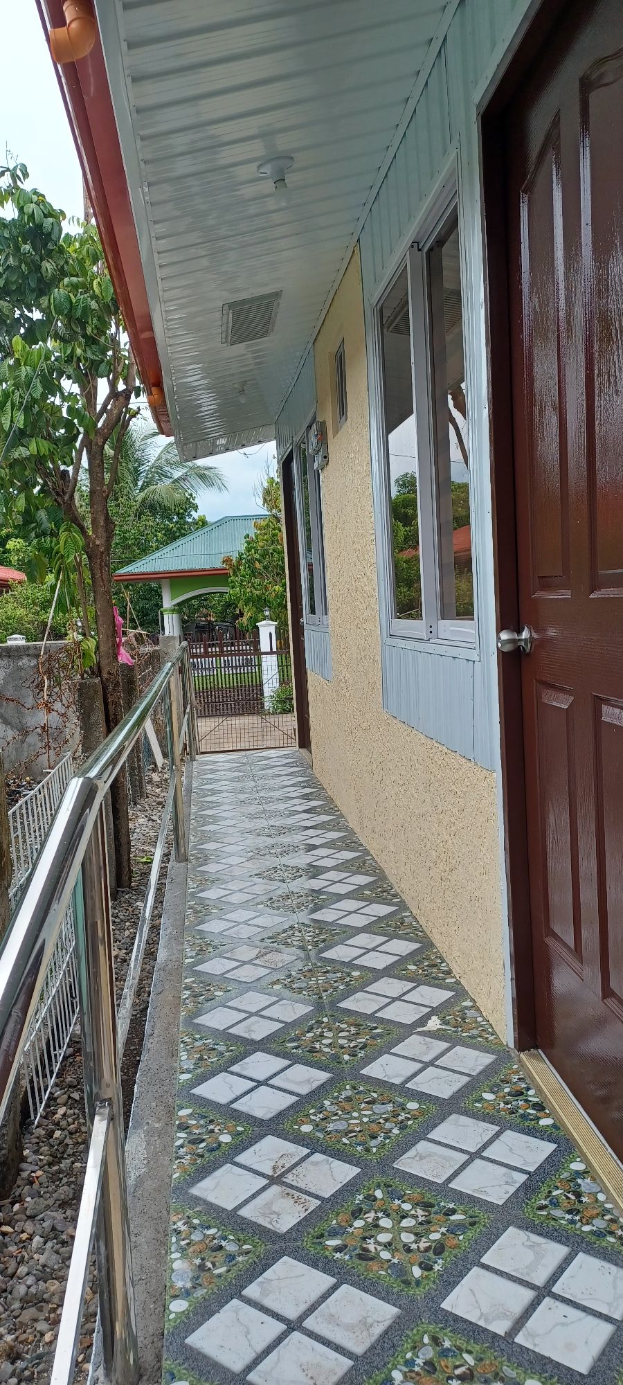 A tiled pathway leads to a brown entrance door, bordered by greenery and visible adjacent structures. The smooth surface of the pathway is complemented by decorative tiles, and natural light illuminates the area, highlighting the inviting atmosphere of the entrance.