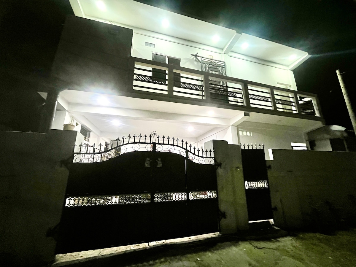The exterior of a modern two-story house is displayed at night, featuring bright lights illuminating the front. A decorative iron gate stands closed, with a surrounding wall providing privacy. The upper balcony is visible, showcasing a railing and additional lighting.