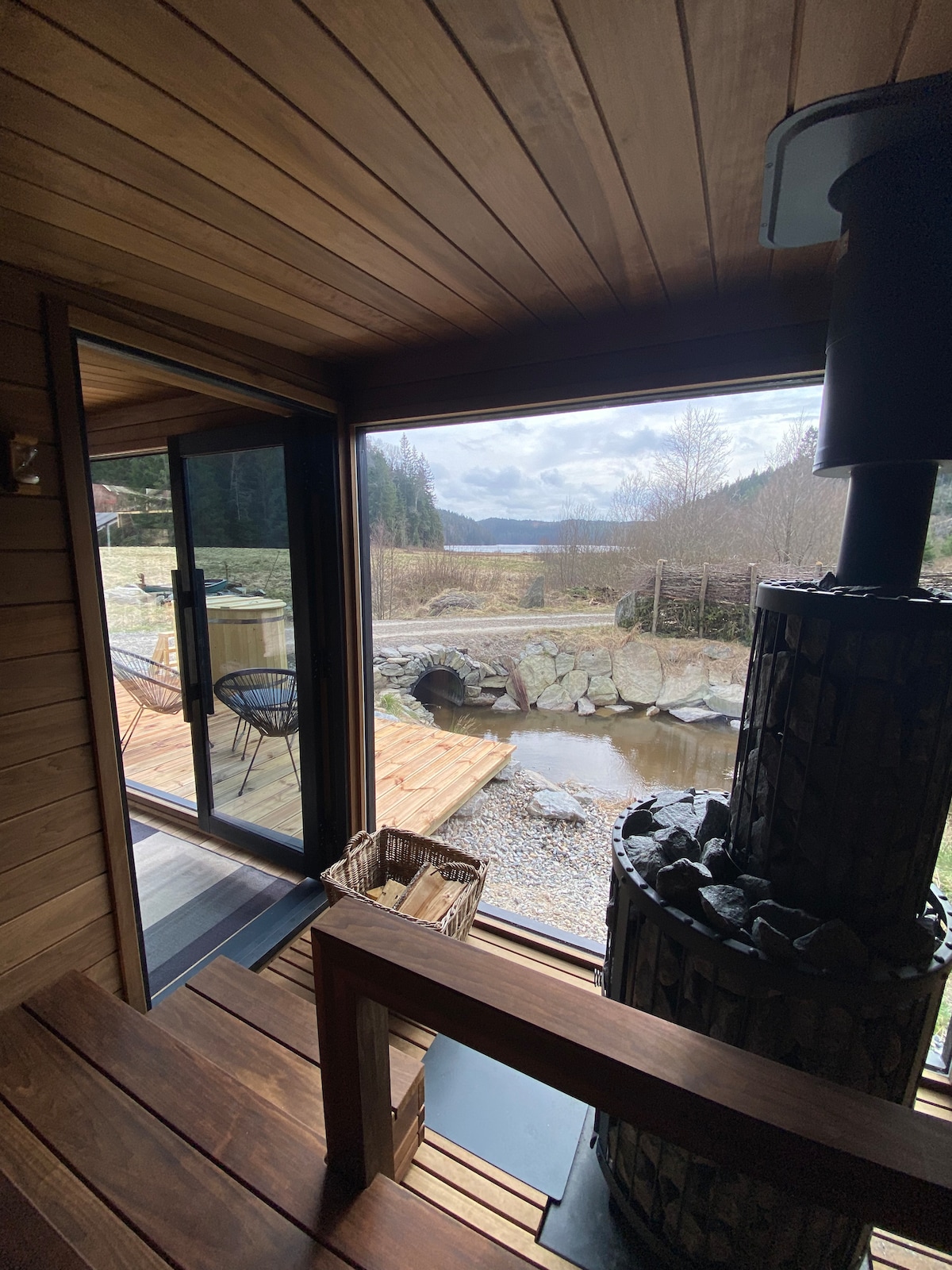 A wooden sauna interior showcases a stone heater beside an inviting wooden step. Large glass doors reveal views of a tranquil outdoor area with a winding stream and lush greenery, promoting a connection with nature.